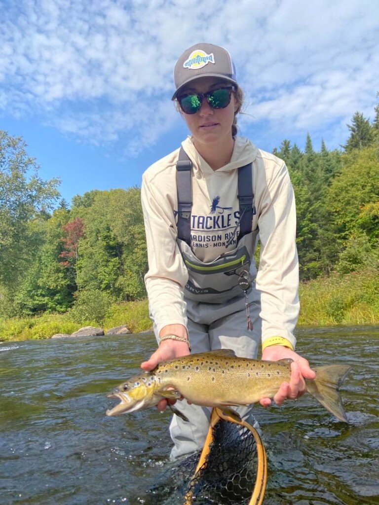 roach 7 A person in sunglasses and a cap holding a large trout over a net in a river, with trees and clear sky in the background during a Maine Fishing adventure.